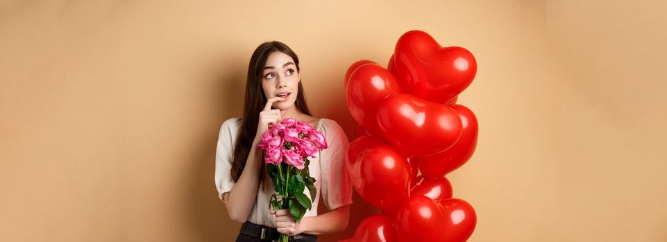 Dreamy young woman holding bouquet of roses and thinking about secret admirer on Valentines day, looking at upper left corner and biting finger, standing near red romantic balloons.