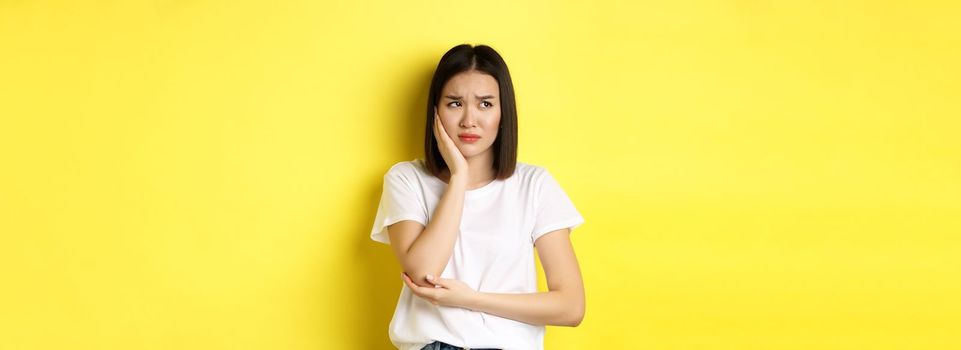 Sad asian woman touching cheek and frowning, having toothache, need see dentist, standing gloomy against yellow background.