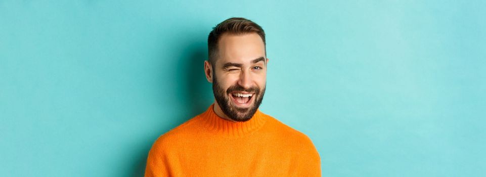 Handsome confident man winking at camera, smiling sassy, standing in orange sweater against blue background.