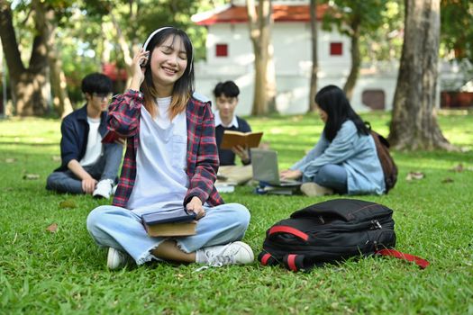 Cheerful asian woman enjoy listening music in headphone while sitting on green grass. Youth lifestyle and education concept.