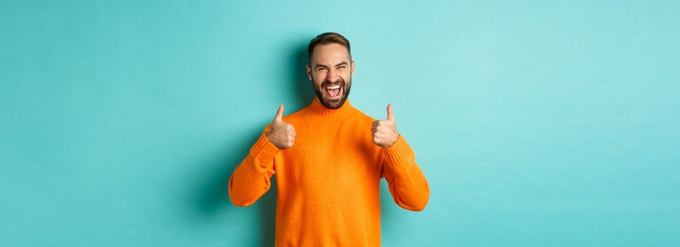 Super good. Satisfied happy man showing thumbs up, agree with you, praise excellent work, looking pleased, standing in orange sweater against light blue background.