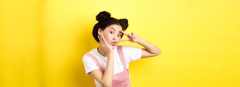 Beautiful asian girl showing v-sign and pouting cute, making silly face with makeup, standing on yellow background.
