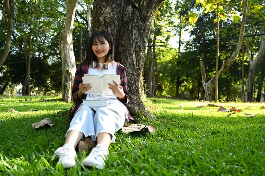 Care free teenage girl relaxing under a tree and reading book on cloudy autumn day. University, school, education concept.