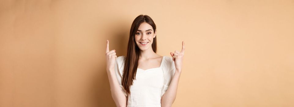 Attractive caucasian woman in dress pointing fingers up, smiling and showing promo banner, standing on beige background.