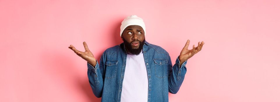 Confused african-american man in beanie looking at upper left corner doubtful, shrugging uncertain, standing over pink background.