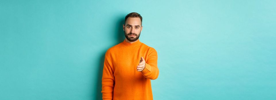 Confident man extending hand for handshake, greeting you, looking self-assured, standing over light blue background. Copy space