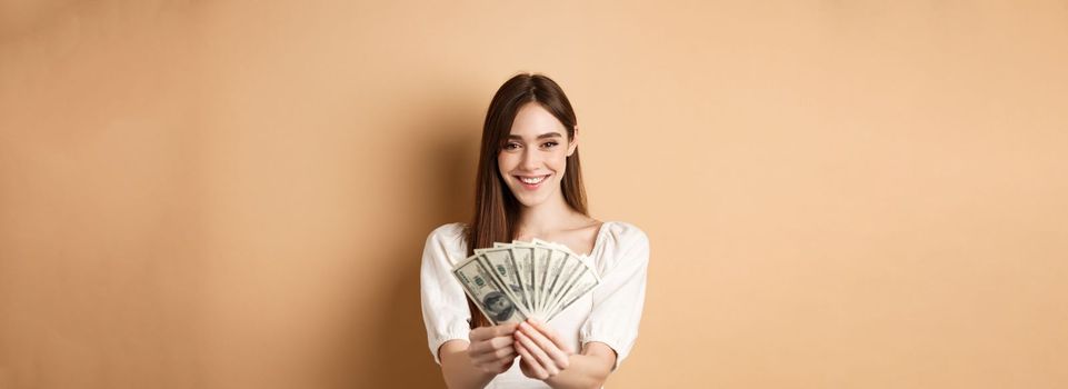 Young woman showing dollar bills and smiling, standing with money on beige background. Concept of loan and insurance.
