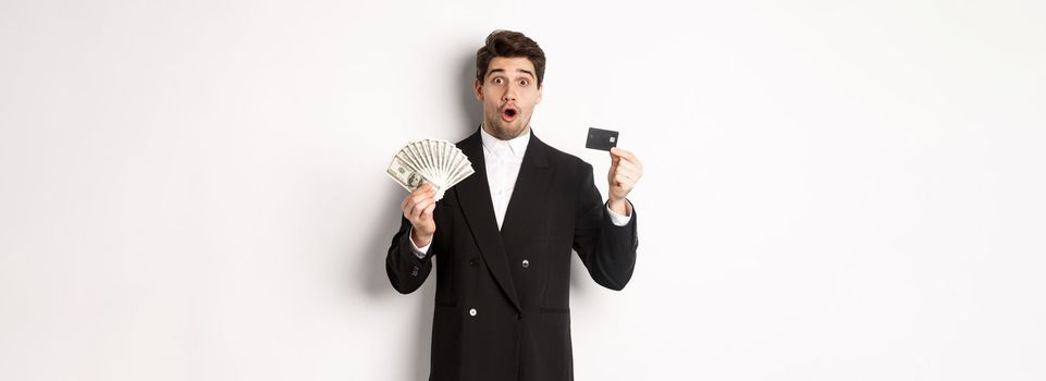 Portrait of surprised handsome man i suit, showing credit card with money, standing against white background.