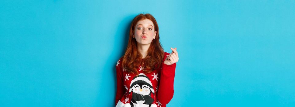 Winter holidays and Christmas Eve concept. Lovely redhead girl in xmas sweater, showing heart sign and pucker lips for kiss, standing over blue background.