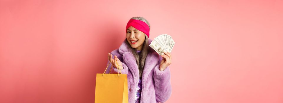 Rich and fashionable asian senior woman wasting money in shops, holding shopping bag and dollars, winking happy at camera, pink background.