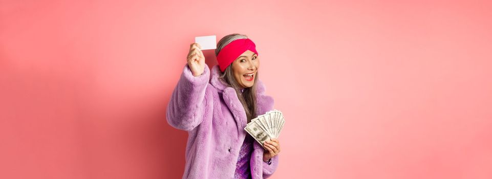 Shopping and fashion concept. Fashionable funky asian woman showing plastic credit card, paying contactless, holding money in other hand, pink background.