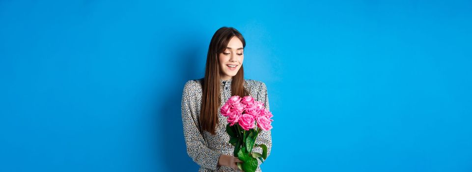 Valentines day concept. Romantic girl in dress looking happy at flowers, smiling at bouquet of pink roses, standing on blue background.
