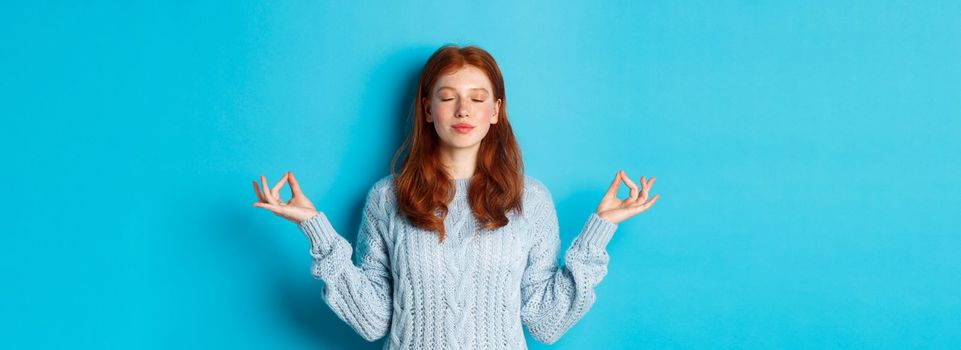 Smiling confident girl with red hair staying patient, holding hands in zen, meditation pose and staring at camera, practice yoga, standing calm against blue background.