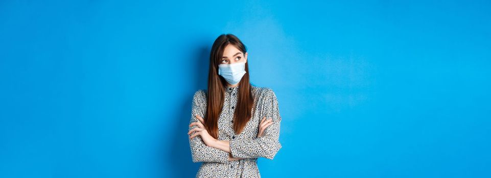 Covid-19, pandemic lifestyle concept. Pensive girl in medical mask look at upper left corner logo, thinking ways of protecting health from coronavirus, blue background.