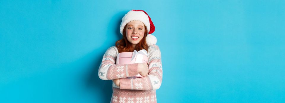 Winter and celebration concept. Dreamy redhead girl in santa hat hugging her christmas gift, smiling happy, standing over blue background.