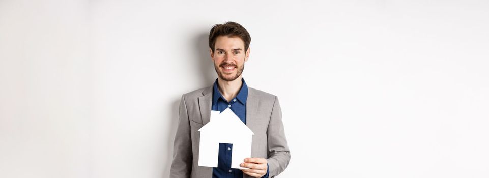 Real estate. Handsome young businessman buying property, holding paper house cutout and smiling, agency advertisement, white background.