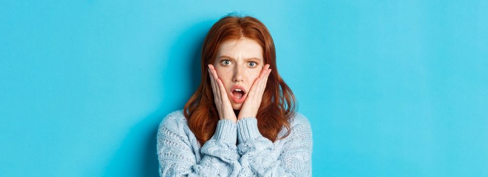 Close-up of shocked redhead girl staring at something displeasing, holding hands on face and gasping, standing over blue background.