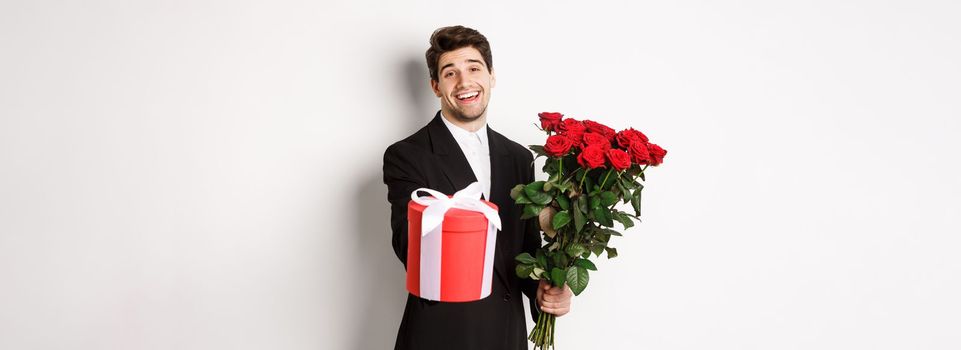 Concept of holidays, relationship and celebration. Image of handsome smiling guy in black suit, holding bouquet of red roses and giving you a gift, standing against white background.