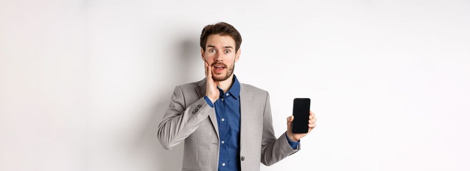 Excited business man in suit showing empty smartphone screen and gasping amazed, demonstrate application, standing on white background.