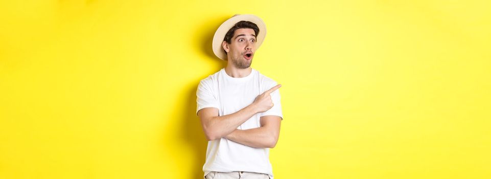 Concept of tourism and lifestyle. Excited handsome guy in straw hat checking out advertisement, pointing and looking at upper left corner logo, yellow background.