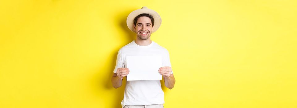 Handsome young male tourist in summer hat smiling, holding blank piece of paper for your sign, standing over yellow background.