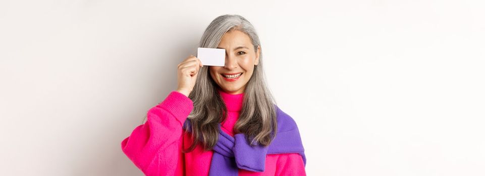 Shopping concept. Stylish asian senior woman smiling and showing plastic credit card, paying contactless, standing over white background.