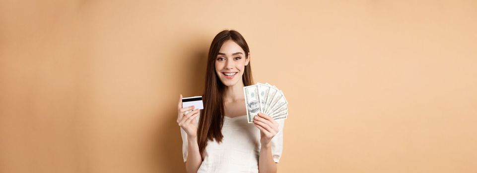 Happy young woman showing dollar bills and plastic credit card, smiling pleased, making money and shopping, standing on beige background.