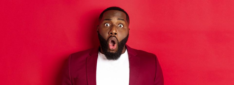 Close-up of shocked Black man gasping and dropping jaw impressed, staring at camera, standing over red background.