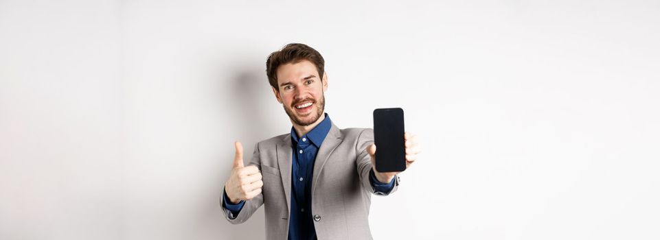 E-commerce and online shopping concept. Cheerful businessman in suit showing thumbs up and empty smartphone screen, recommending app, standing on white background.