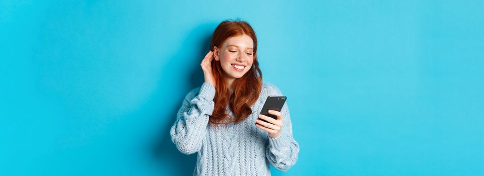 Technology. Happy redhead girl looking pleased at mobile phone, reading compliment in message, smiling and tuck hair behind ear, standing in sweater over blue background.