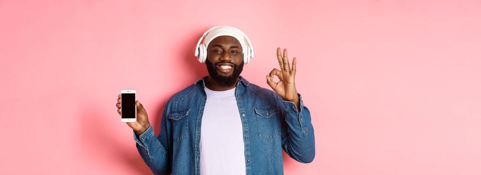 Happy hipster guy listening music on headphones and showing mobile screen, smiling satisfied, showing okay sign in approval, like playlist, standing over pink background.