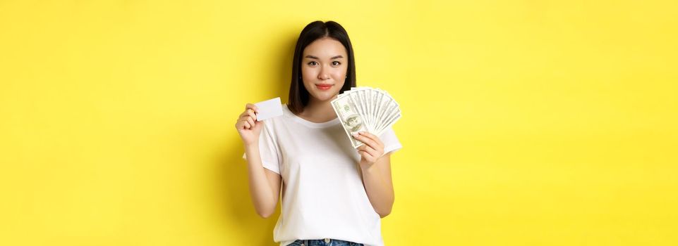 Beautiful asian woman with short dark hair, wearing white t-shirt, showing money in dollars and plastic credit card, standing over yellow background.