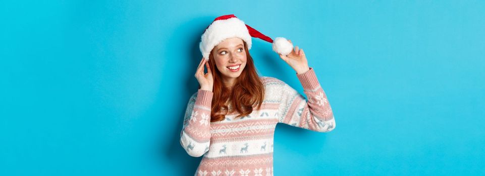 Winter and Christmas Eve concept. Cute teenage girl wearing santa hat and sweater, celebrating xmas, smiling happy, standing over blue background.