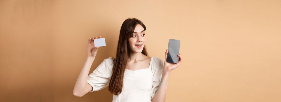 Check this out. Smiling happy young woman showing plastic credit card and empty mobile phone screen, demonstrate application, standing on beige background.