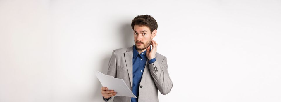Confused office worker in suit scratching beard and look clueless, cant understand document, holding paper with indecisive face, white background.