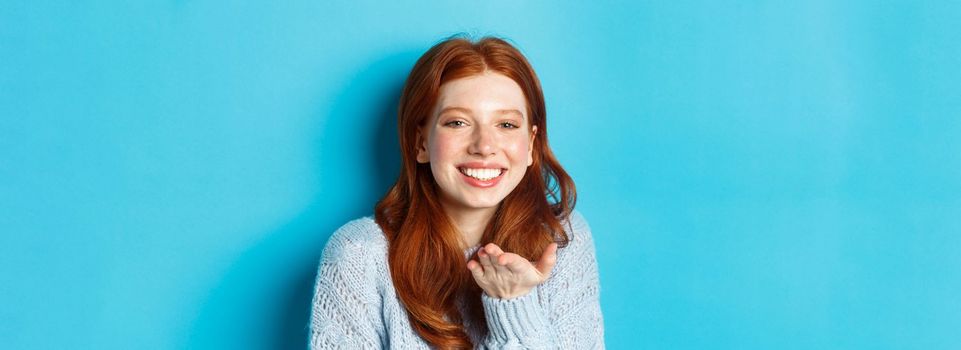 Lovely redhead female model smiling, sending air kiss at camera, standing against blue background.