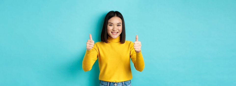 Cheerful asian female model showing thumbs up gesture, smiling and looking impressed, praise good product, standing over blue background.