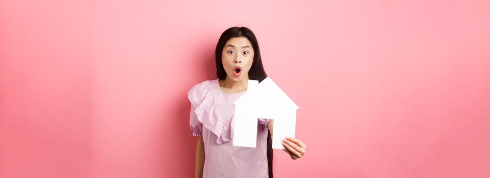 Real estate and insurance concept. Excited asian woman showing paper house cutout and look amazed, special offer, standing on pink background.