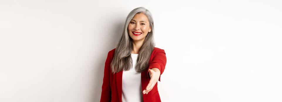 Professional asian businesswoman with grey hair, saying hello, stretch out hand for handshake and smiling, standing over white background.