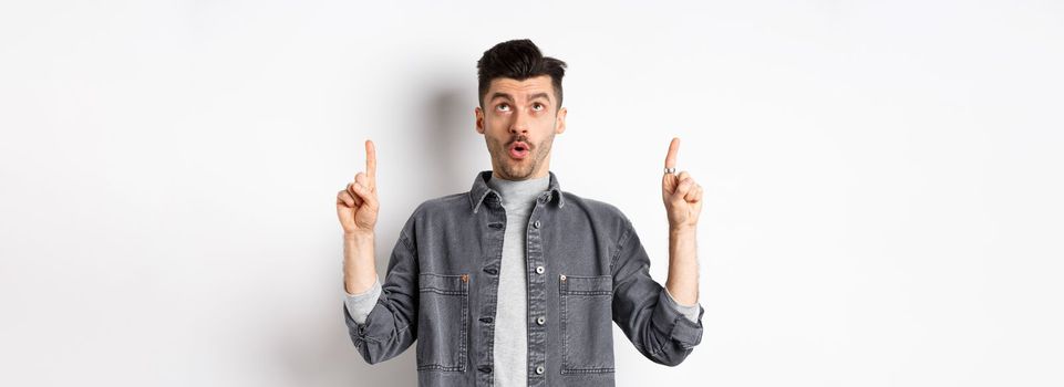 Excited modern man in denim jacket looking and pointing up, gasping amazed, checking out cool offer, standing on white background.