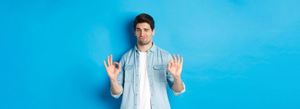 Satisfied handsome man showing a-ok signs and looking pleased, approving something good, standing against blue background.