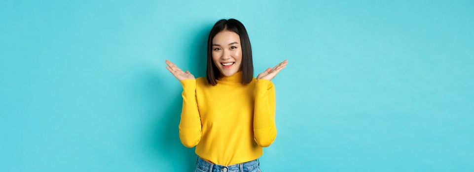 Beauty and fashion concept. Attractive japanese girl raise hands up and demonstrate something, smiling happy and looking at camera, showing promo, blue background.