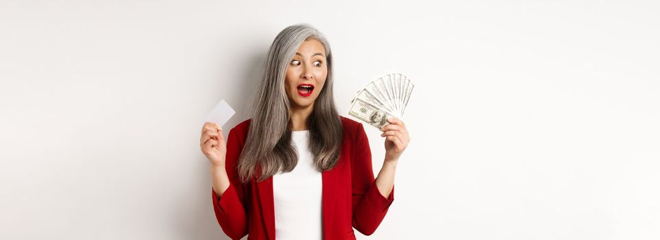 Surprised asian middle-aged woman with grey hair, looking at money and holding credit card, earning cash, standing over white background.