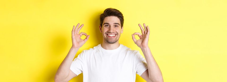 Close-up of handsome young man showing okay sign, approve and agree, smiling satisfied, standing over yellow background.