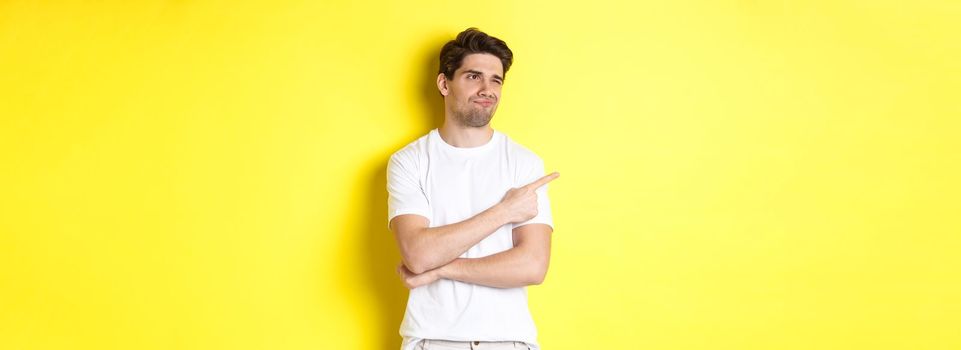 Unimpressed young man complaining, pointing and looking with skeptical face at bad product, standing against yellow background.