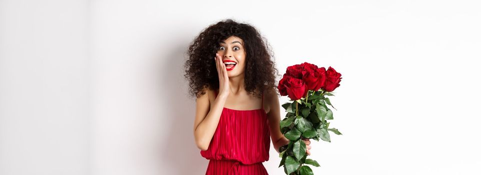 Excited curly woman in red dress, receive bouquet of roses and look surprise, rejoicing from romantic gift, standing on white background.