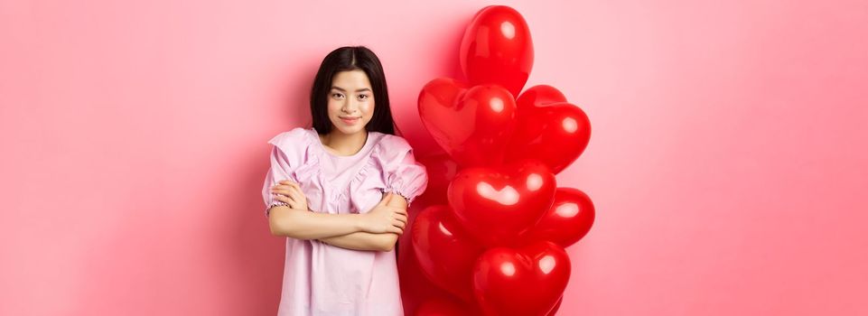 Confident teenage girl cross arms on chest and smile, celebrating valentines day in cute dress with red heart balloons, standing on pink background.