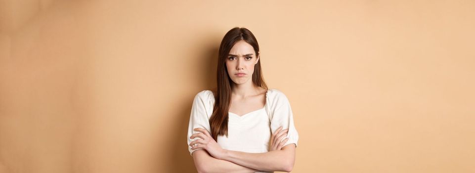 Angry girlfriend cross arms on chest and frowning, looking offended or disappointed, sulking at you, standing mad on beige background.