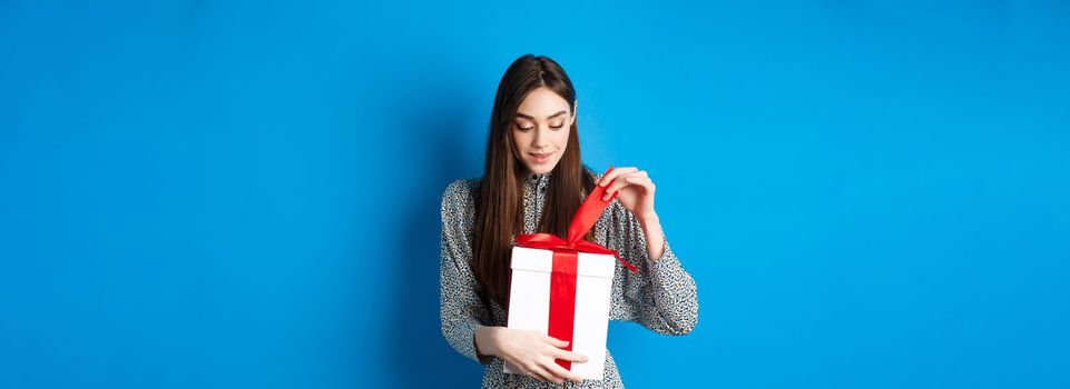 Valentines day. Cute young woman open box with gift, take-off ribbon from present and smiling intrigued, standing on blue background.