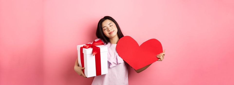 Valentines day concept. Romantic and tender asian teen girl smiling, close eyes and standing dreamy with lover gifts, holding present and big red heart cutout, pink background.